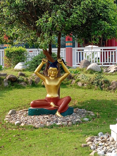 The image captures a golden statue of a figure in a seated position with arms raised, as if tying hair. The statue is located in Lumbini Sanskritik, Nepal, in a garden-like setting with a lush green lawn, scattered rocks, small shrubs, and a tree under which the statue sits. The statue rests on a dark green base, surrounded by small stones. A white fence with red and ornate pillars is visible in the background, along with greenery and what appears to be a building or structure. The lighting suggests it is daytime.