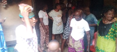 In a candid indoor shot from Ohafia, Nigeria, several individuals are gathered. On the far left, a woman with a serious expression carries a green basin on her head, containing a clear plastic package of what appears to be food or goods. She wears a light-colored top and patterned bottoms. To her right, another woman in a light green, short-sleeved dress featuring a large Hello Kitty design stands with her hands on her hips. Next to her, a man in a white T-shirt with a "50" or "50th" logo on the chest is visible, wearing patterned trousers. To his right, another woman in a vibrant green and yellow patterned traditional outfit is partially seen. The head of a small child is visible at the bottom center foreground. The background is dimly lit and indistinct, suggesting an indoor setting or a shaded area. The overall scene depicts a casual gathering or an everyday interaction.