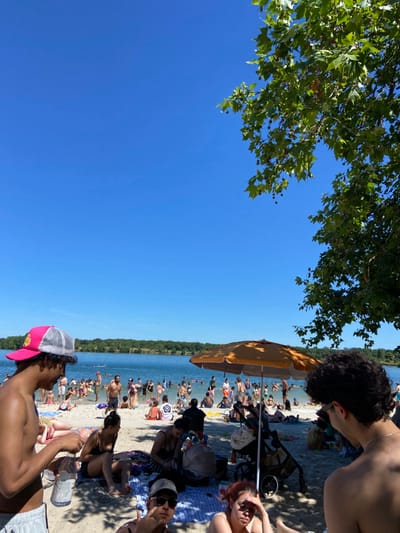 The image shows a scene of people enjoying a sunny day at a lake in Tournefeuille, France. The coordinates provided, latitude 43.568 and longitude 1.344, correspond to a location near the Lac de la Ramée. The image shows numerous people on the sandy shore and in the water of the lake. There is a large, orange umbrella in the center of the image, providing shade for several people. The foreground shows a man with a pink baseball cap, facing the camera and holding a water bottle, interacting with another man wearing a white shirt. A woman in a bathing suit is sitting in front of him, while another woman is further back, to the right.  To the right, a man with curly hair is standing with his back to the camera, looking to the right.  The background shows a large group of people enjoying the lake, some wading in the water, some sitting on the shore, and others walking along the edge of the lake. The image captures a relaxed and enjoyable atmosphere at the lake. The sky is bright blue and clear, indicating a sunny day. The trees in the background and in the upper right corner of the image provide shade, creating a contrast between the bright sun and shaded areas.  There is a dense growth of green vegetation at the edge of the lake, which further enhances the beauty of the scene. The image captures a typical summer day, with people enjoying the outdoors.