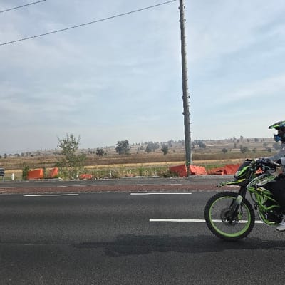 A motorcycle speeds along a black asphalt road, passing a utility pole. The rider, wearing a helmet, is partially visible. The motorcycle is green with lime green rims. The roadside is lined with orange barriers, beyond which is a field of brown and green vegetation. The sky is overcast, creating a hazy atmosphere. Distant trees dot the landscape.