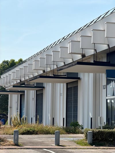 The image shows the exterior of a modern building with a distinctive roofline. The building is white with vertical ribbing. The roof is a sloping structure covered with what appears to be glass panels, interspersed with angled, white opaque elements that cast shadows. There are multiple covered entrances along the building's facade, marked with numbers such as "10" and "100". The entrances themselves are covered by dark, projecting awnings and have metal grate doors. In front of the building, there is an area with grey concrete bollards and an asphalt surface. To the left of the building, overgrown vegetation and tall, dried grasses are visible. The sky above is clear and blue. The overall scene is one of a commercial or industrial building in a bright, sunny environment. Based on the "Rungis" signage visible on the building in the upper right quadrant, this location is identified as being in the city of Rungis, France.