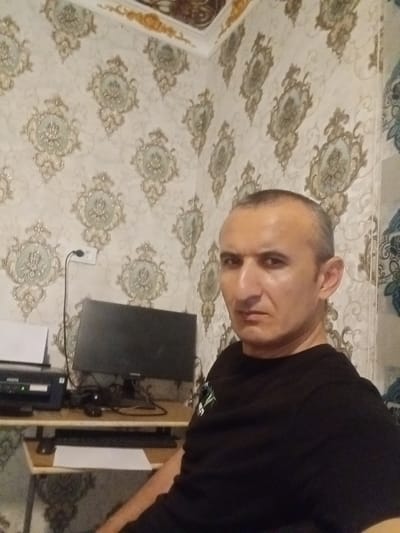 A man with short, dark hair and a serious expression is seated in front of a desk in a room. He is wearing a black t-shirt. On the desk are a computer monitor, a printer, and a keyboard. The walls are covered with a patterned wallpaper featuring ornate medallions. There is also an electrical outlet visible on the wall. The image appears to be a selfie taken indoors. The location is identified as the city of Karagana, Uzbekistan.