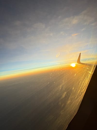 The image is a photograph taken from the window of an airplane. The sun is setting, casting a warm golden light across the scene. A rainbow is visible, arcing across the sky, its colors vibrant against the clouds. The wing of the airplane is also visible, jutting out into the frame. The sky is a mix of blues and grays, with some wispy clouds. The overall impression is one of beauty and tranquility. The time of day is sunset, and the weather appears to be clear with some clouds. The location is implied to be in the sky, above Huacho, Peru, though no specific landmarks are visible. The emotion evoked is one of awe and wonder at the natural beauty of the sunset and rainbow. There is no visible text in the image.