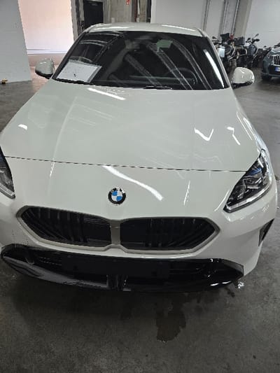 A white BMW car is parked indoors, presented in a frontal, slightly elevated perspective. The vehicle, featuring a prominent black kidney grille with vertical slats and a central BMW emblem, appears to be a modern model. Its headlights incorporate blue-tinted LED elements. A glossy black lower bumper/splitter is integrated below the main grille. A white document or tag is visible on the dashboard, placed against the windshield from the interior. The car is situated on a grey concrete floor, which exhibits a dark, irregularly shaped liquid stain directly beneath the front bumper. The background on the left consists of a plain white wall with a dark doorway opening and a small yellow object at floor level. To the right, a line of motorcycles, primarily identifiable as BMW GS models, is parked. A dark-colored vehicle is partially visible beyond the motorcycles. Overhead lighting is reflected in the car's pristine, white bodywork and windshield. No individuals are present in the scene. The location is an indoor facility, likely a dealership showroom or service area, in Sprendlingen, Germany.