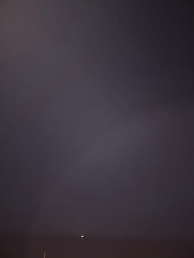 The image depicts a dark, cloudy sky over Platja D'aro, Spain. A faint band of light, possibly from distant city lights or a slight break in the clouds, is visible in the lower portion of the frame. A few small, bright specks, likely stars or distant lights, are scattered across the sky. The overall scene suggests a night or twilight setting with limited visibility due to atmospheric conditions.