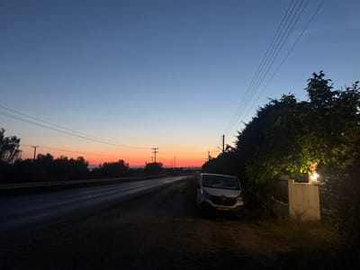 The image depicts a road running parallel to a coastline at twilight in Γερακινή, Greece. A white van is parked on the roadside next to a tree and a fence with a lit lamp. Power lines are overhead. The sky displays a gradient of colors from dark blue to orange near the horizon. Trees and vegetation line both sides of the road.