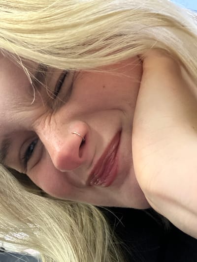 An extreme close-up shows a woman's face tilted at an angle. She has blonde hair, pale skin, and a nose ring. One eye is slightly open, showing a blue iris. The other eye is squinting. Fine lines and wrinkles are visible around the eyes. Her lips are glossy and partially open. Part of her arm or hand is visible above her head. The background is dark.
