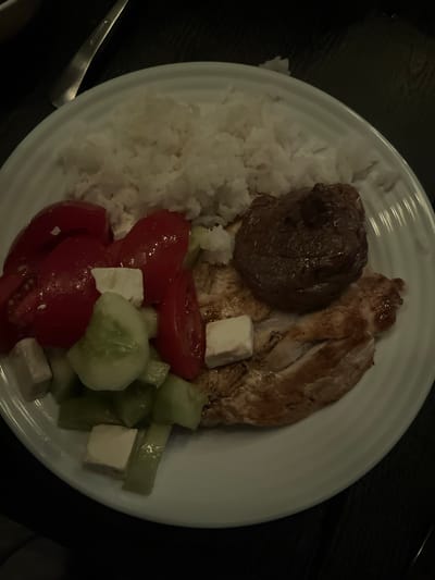 This image shows a close-up view of a meal served on a white plate. The meal consists of several components: a mound of white rice, a piece of dark, possibly beef, meat, and grilled chicken breast. Alongside the meats and rice, there is a salad made with chopped tomatoes, cucumbers, and cubes of white cheese, likely feta. The overall presentation suggests a home-cooked meal or a casual dining experience. The lighting in the image is dim, which could indicate it was taken in the evening or indoors. There are no visible people in the frame, nor any explicit text. The dish appears to be a balanced meal with protein, carbohydrates, and fresh vegetables. The presence of feta cheese and the general composition of the meal might hint at Mediterranean or Greek cuisine, aligning with the provided location context of Chaidefto, Greece.