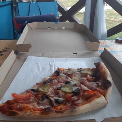The image shows a partially eaten slice of pizza inside a cardboard pizza box. The pizza has mushrooms, pickles, and tomato slices on it. The box is open and sits on a wooden table, with a blurry background of greenery and wooden structures. To the left of the box, a blue chair with a dark blue rolled-up item is visible. The lighting suggests it is daytime. The text "dance" is partially visible on the side of a wooden structure below the pizza box.