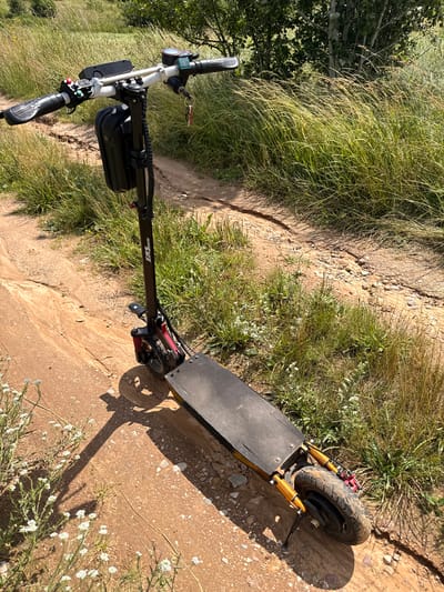 An electric scooter is parked on a dirt path in a grassy, rural area. The scooter is black and red with a black bag attached to the handlebars. The path is a light brown dirt with scattered small rocks. Tall green and yellow grass flanks the path, with some trees visible in the background. The scene appears to be outdoors on a sunny day. The scooter's brand name, "BUNSUN," is visible on the handlebar stem.