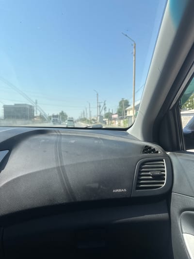 The image shows the interior of a car, likely in Ынтымак, Kyrgyzstan, looking out through the windshield. The dashboard is visible on the right, with a textured surface and an air vent. The word "AIRBAG" is printed on the dashboard. The windshield is somewhat dusty, with a few smudges and scratches.

Through the windshield, the scene outside appears to be a street with buildings and trees. There are utility poles and wires visible, suggesting an urban or suburban environment. The sky is clear and blue, indicating it is daytime with sunny weather. The orientation of the image suggests it was taken from the driver's or passenger's seat. No people are visible in the image.