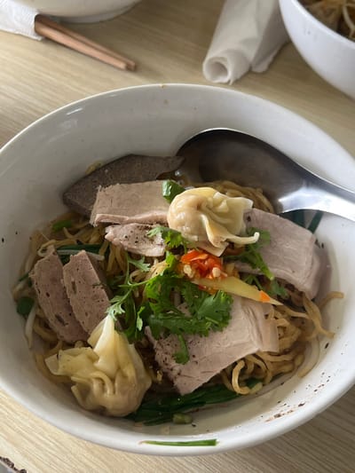 A close-up shows a bowl of noodles, likely Asian cuisine. The noodles are topped with slices of pork, liver, Vietnamese ham, and dumplings. Garnishes include fresh coriander, chilies, and other herbs. A large spoon rests in the bowl. The dish is served in a white bowl and appears to be on a light-colored wooden table, with chopsticks and another bowl partially visible in the background.