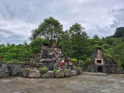 A cloudy, overcast day frames a scene of a rock garden with a waterfall feature and a stone barbecue pit in Charnali, Georgia. The waterfall is constructed from a variety of stones, with greenery and potted plants interspersed throughout. A red pipe is visible on the waterfall structure. To the right, a substantial stone barbecue pit stands with an arched opening at its base and a chimney atop. The ground in front is paved with concrete, showing signs of recent moisture. Lush green trees and vegetation form a dense backdrop to the man-made elements, with a hillside visible in the distance. There are no people or animals visible in the image.