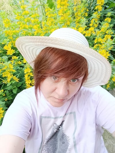 A selfie is taken by a person wearing a straw hat and a light purple t-shirt. The person has auburn hair and light-colored eyes. They are looking directly at the camera with a neutral expression. The background is filled with bright yellow flowers and green foliage. The lighting suggests it is daytime. The location is identified as Вережи Снт, Russia. The t-shirt has a graphic of a grey animal, possibly a cat, printed on the front. No visible text is present on the clothing or in the background. The image captures a moment of casual outdoor activity, possibly in a garden or natural setting.