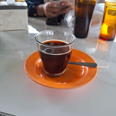 A close-up, eye-level shot captures a glass of dark coffee resting on an orange saucer. The coffee has a thin layer of foam around the rim. A spoon with a decorative pattern rests on the saucer to the right of the glass. In the background, out of focus, a person is visible holding a smartphone. To the right of the coffee, two amber-colored bottles are partially visible, one darker than the other. The scene appears to be set on a table with a marbled surface, likely in a cafe or restaurant. The lighting suggests an indoor setting with natural light contributing to the reflections on the table and bottles. The location in Mesjid Tuha, Indonesia is inferred from the provided context, but not visually evident in the image itself.