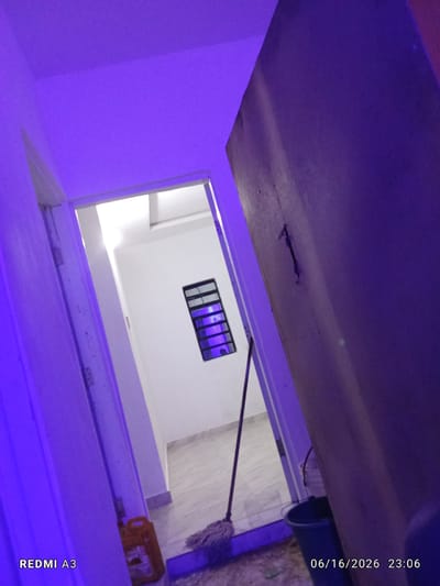 The image shows an interior scene, possibly a hallway or entryway. The walls and ceiling are illuminated with a strong purple light, creating a dramatic and somewhat surreal atmosphere. To the left, a doorframe is visible, painted white and showing signs of wear. Through an open doorway ahead, a brightly lit room with white walls can be seen. A small, rectangular window with dark panes is mounted high on the far wall of this room. A mop with a light-colored head rests against the wall, with its handle extending upwards and out of frame. To the right of the doorway, a large, dark-colored door dominates the foreground, obscuring much of the right side of the image. A yellow plastic container is visible on the floor near the doorway, and a dark blue bucket stands near the large door. The floor appears to be tiled. The timestamp in the bottom right corner indicates the date as 06/16/2026 and the time as 23:06. The text "REDMI A3" is visible in the bottom left corner. The overall impression is one of a functional space, perhaps a home or business, illuminated by colored lighting. There are no people visible in the image.