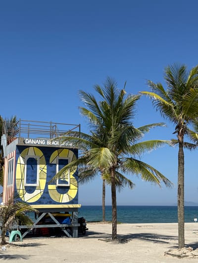 The image shows a lifeguard station on a sunny beach in Da Nang, Vietnam. The station is painted blue with yellow numbers "06" and white windows. Above the station, "DANANG BEACH LIFECUBE" is written. Palm trees line the beach, which has a sandy surface and leads to the calm, blue ocean under a clear blue sky. A red and white striped section is visible on the left side of the lifeguard station. Beneath the station, there are some objects stored, including life vests and what appears to be a motorcycle on the right. The overall environment is serene and depicts a typical beach scene.