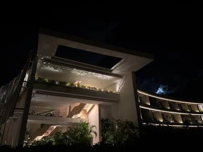A modern building is shown at night. It features layered levels with open spaces and staircases. Green plants are visible on some levels. The design incorporates a rectangular opening on the top level. To the right is a curved building with illuminated balconies. The background is a dark sky with the moon peeking through the clouds. Shadows and light interplay create a visually interesting architectural scene.