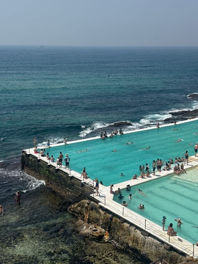 An aerial view captures a pool filled with people, adjacent to the ocean. Waves crash against the pool's edge and the surrounding rocks. Swimmers and sunbathers populate the pool area. The ocean stretches out to the horizon under a hazy sky. A bird soars over the water's surface. The pool appears to be constructed on a rocky outcrop along the coastline.