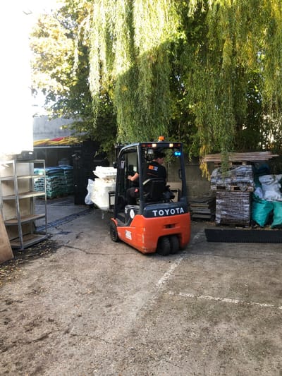 This image depicts an outdoor material storage or industrial yard in Houilles, France.

A male individual is operating an orange and black Toyota counterbalanced forklift, positioned roughly in the center-right of the frame, facing slightly left. He is seated in the driver's compartment, wearing a black short-sleeved shirt with an orange logo on the left chest. The forklift has an illuminated amber rotating beacon on its upper left and a blue warning light on its upper right. It is actively carrying a stack of several large, rectangular bundles of white material, possibly wrapped in clear plastic, on its forks.

To the left, a metal shelving unit with four empty shelves is visible in the foreground, with a sheet of plywood leaning against it. Further back on the left, stacks of dark-colored bags, possibly on pallets, are arranged, along with stacked black plastic crates and some yellow and red pipes under a partial shelter.

The right side of the image features a large tree with dense, weeping green foliage, casting shadows across the scene. In the midground on the right, stacks of wooden pallets are visible, alongside green bags and other bundled items. A black, grid-like mat or panel lies on the ground in the lower right corner.

The ground is an uneven, light-colored concrete or asphalt surface with cracks and scattered debris, including leaves. A faded white line is present on the ground in the lower right. Strong sunlight illuminates the scene, creating distinct shadows. A light-colored building wall is partially visible on the far left.