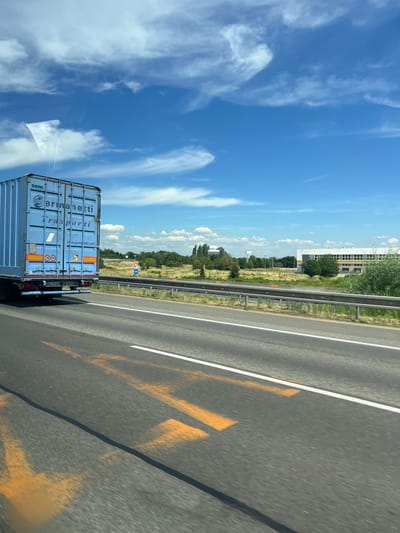 The image shows a blue cargo container truck on a highway under a blue sky with scattered clouds. The truck has "Germanetti Trasporti" written on its side. Orange markings are visible on the road. In the background, there is a grassy field, trees, and buildings. A speed limit sign indicating "50" is also visible. The scene appears to be a sunny day on a highway in or near Senec, Slovakia.
