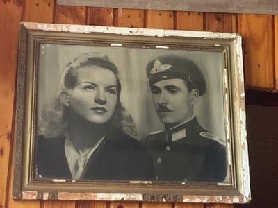 This is a black and white studio portrait of a man and a woman, framed in an ornate, weathered frame. The woman is positioned on the left, with her head slightly turned to the right. She has wavy hair and is wearing a dark jacket and a light-colored scarf or tie. The man is on the right, wearing a military uniform that includes a cap with an emblem, a jacket with epaulets, and a mustache. He is looking slightly to his left. The background appears to be a studio backdrop, possibly a curtain or draped fabric. The lighting is soft, typical of portrait photography from an earlier era. The overall tone of the image is formal and serious, with both subjects appearing composed. The frame itself shows signs of age, with chipped paint and a distressed appearance, suggesting the photograph is an heirloom. The photo is hanging on a wooden wall, with visible wood grain and some imperfections in the wall surface. Based on the clothing and photographic style, the portrait likely dates from the mid-20th century. The location context of Novo bardo, Bulgaria, suggests this portrait might be of individuals with ties to that region. There is no visible text in the image.