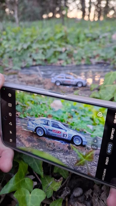 The image shows a person holding a smartphone, using it to take a picture of a toy car. The toy car is placed on a surface covered with dirt, leaves, and vegetation. The scene appears to be outdoors, potentially in a garden or park-like setting, and the background is blurred, but appears to be an urban environment. The picture is taken in the city of Podbadac, Montenegro.