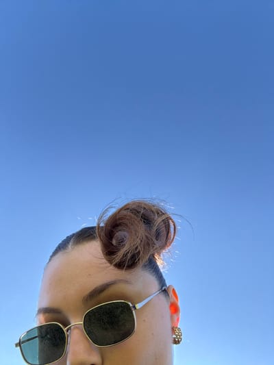 Looking up at a person against a bright blue sky, the perspective is from below. The person's hair is styled in a bun, and they wear sunglasses with gold frames. An orange scarf and a chunky gold ring are visible near the ear. The person's face is partially visible, showing an eyebrow and the bridge of their nose.