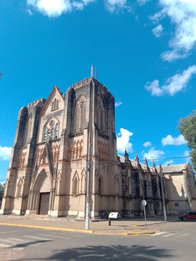 This image captures a low-angle, wide shot of a large, stone church with Gothic architectural elements, set against a bright blue sky dotted with white clouds. The church has a prominent central tower and spires, with arched windows and intricate stone detailing. The facade is a light beige or tan color, showing signs of age and weathering. A large, dark wooden double door is visible at the main entrance. The church is situated in a city environment, with a paved street and a crosswalk in the foreground. A yellow curb lines the street. A single red car is parked on the right side of the street. A tall, thin utility pole stands to the right of the church. The environment suggests a sunny day in a Brazilian city, possibly Cáceres, Brazil, given the architecture and clear skies. There are no people visible in the image.