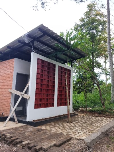 The image captures a small, single-story building in Moe Gyoe, Myanmar, situated within a heavily vegetated area under an overcast sky. The structure features a corrugated metal roof on a dark metal frame, with a light fixture visible beneath the eaves. The building's left side is constructed from red-brown brick masonry. Its front facade displays a white-painted wall with two prominent, rectangular sections filled with dark red/maroon decorative ventilation blocks. A dark-colored door is present on the left side of the building, partially obscured by an 'X'-shaped wooden brace leaning against the wall. An electrical cable extends towards the roof from the upper left. The immediate foreground shows an area partially paved with light-colored rectangular pavers, with loose pavers stacked on bare earth to the left. A slender, young tree with green foliage grows directly in front of the building from the paved section. The background consists of dense green foliage and numerous tall trees. No individuals are present.