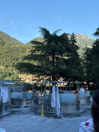 The media file depicts an outdoor thermal spa with several circular, clear-walled pools filled with bubbling water. Several people are seen relaxing in the pools, some conversing while others soak. The setting is mountainous, with lush green hills and trees forming a backdrop. A building with orange facades is visible further up the hillside, suggesting a resort or hotel. Large evergreen trees, possibly cedars, are prominent in the foreground, their branches creating a natural canopy. The pools are surrounded by a paved area, and white robes are hanging on racks near some of the pools, indicating the nature of the establishment. A yellow warning sign with black text and a symbol of a person slipping is visible on the right side, cautioning about wet floors. The sky is a clear, bright blue, and the sunlight suggests it is daytime, likely late afternoon or early evening given the long shadows. The overall atmosphere appears tranquil and leisurely.