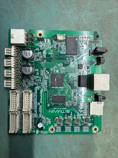 The image displays a green printed circuit board (PCB) with various electronic components. The PCB is laid flat on a textured, greenish-gray surface, suggestive of a workbench or industrial setting. The board is oriented with its longer side running horizontally across the frame.

Prominent components include several white connector blocks with multiple pins, arranged in rows along the left and bottom edges of the PCB. There are also two cylindrical silver capacitors with black tops, each labeled "330 35V VT". A large black integrated circuit (IC) with text that appears to read "1487CC" is centrally located, with another smaller black IC nearby. A prominent white rectangular connector with pins is situated at the top left. There are also several smaller black components resembling surface-mount ICs and resistors.

Towards the right side of the PCB, there are two metallic-looking connectors with square openings, one larger than the other. A small red LED is visible near the top right corner. Text printed on the PCB is visible in several areas, including "BITMAIN" in large white letters near the bottom center, and what appears to be a model or serial number starting with "10000579" near the bottom left. There are also many smaller alphanumeric labels identifying components (e.g., R102, C179, U2, J11).

The overall impression is that of a piece of electronic hardware, possibly a control board or module, photographed for identification or inspection.

This image does not contain any visual information that would allow for the determination of its location in Indiantown, United States of America.