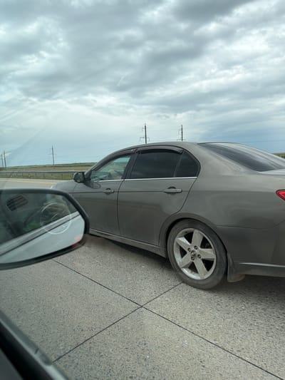 A medium shot shows the side of a gray four-door sedan driving on a highway, possibly near Damsa, Kazakhstan, as indicated by the distant, sparsely populated landscape and the presence of power lines stretching across the horizon. The car's windows are tinted, and a driver is partially visible behind the steering wheel, appearing to be wearing light-colored pants. The vehicle has a weathered appearance, with dust and dirt coating its lower body and wheels. The front passenger side wheel is prominently featured, revealing a five-spoke alloy rim that shows signs of wear and tear. A side-view mirror reflects the interior of another vehicle, suggesting the photo was taken from the passenger seat of a car traveling alongside the sedan. The road is paved with concrete, marked by lines indicating lanes. The sky above is overcast with heavy, gray clouds.