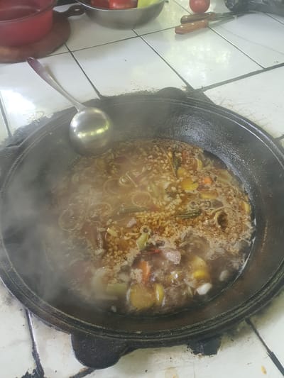A black, cast-iron pot filled with a steaming stew is the central subject of the image. The stew appears to be a pilaf or similar dish, containing rice, meat, carrots, potatoes, onions, and other vegetables. A silver ladle rests inside the pot, positioned to the left of the frame. The pot is placed on a tiled surface, likely a stovetop, as indicated by the grout lines between the white tiles. To the left of the pot, a red tomato and two wooden-handled knives are visible, suggesting ingredients for the meal. A partially visible metal bowl containing what appears to be more produce is also present. The scene is illuminated by natural light, suggesting it could be daytime. The presence of steam rising from the pot indicates the food is actively cooking or has just been served. The overall setting appears to be a kitchen or an outdoor cooking area in a warm climate, given the type of food and preparation. There is no visible text in the image.