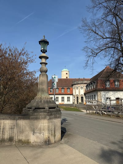 The image displays an exterior scene of the Sprudelhof complex in Bad Nauheim, Germany, captured on a sunny day under a clear blue sky with two faint contrails.
In the immediate foreground, to the left, a tall, ornate stone lamp post with a dark, possibly bronze-green, lantern housing stands atop a substantial, light-grey stone structure, likely a bridge railing. The lamp post base features carved details, and the supporting wall below contains a rectangular access panel. Adjacent to the left, a leafless deciduous tree with visible catkins is present.
An asphalt road occupies the center-right foreground, curving towards the background. To the right of the road, a wooden fence structure runs parallel to the asphalt. Behind this fence, a silver sedan is partially parked. A bare deciduous tree frames the upper right portion of the image.
In the midground, two vertically stacked white square signs, each with a green border and a black bicycle pictogram, are visible. The upper sign displays a left-pointing arrow, and the lower sign indicates a straight direction.
The background is dominated by the historic Sprudelhof complex. This multi-building structure features red-tiled roofs, numerous white-framed dormer windows, and light-colored facades. A prominent, cylindrical, light-colored tower with a conical turquoise-green roof is positioned on the left side of the complex. The central section exhibits a circular, neoclassical architectural element with large windows. The ground in front of the buildings is a combination of paved surfaces and green grass patches. No human figures or animals are visible.