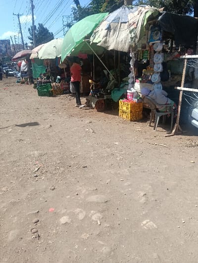 This image depicts an outdoor scene of multiple informal vendor stalls situated along a dirt path or roadside in Ndenderu ward, Kenya, under bright daylight. The ground is dry, unpaved earth with scattered small rocks, minimal vegetation, and visible footprints, including a distinct impression in the foreground.
To the left, several structures are visible, including power lines, utility poles, distant vehicles, and trees under a clear sky. A row of at least five makeshift stalls extends from the middle-ground towards the right. These stalls are constructed with wooden frames and covered by various umbrellas and tarpaulins for shade, including a prominent green umbrella bearing the "Safaricom" logo and a multi-colored umbrella.
Approximately three individuals are visible. A person wearing a red/orange shirt and dark trousers stands facing right, appearing to interact with a seated individual at one of the central stalls. This seated person, partially obscured, is dressed in light-colored clothing. Further to the right, another individual is seated on a grey plastic chair under a separate stall, covered in light-colored fabric or a blanket.
The stalls display various goods. Green plastic crates filled with produce, including bananas, are visible. Yellow plastic crates are stacked at the right-most stall, containing various items. Other merchandise, such as packaged goods, white circular items, and what appear to be household products, hang from the stall structures or are arranged on low surfaces. A wooden frame with empty clothes hangers is present at the far right. The overall environment suggests a bustling, informal market area.