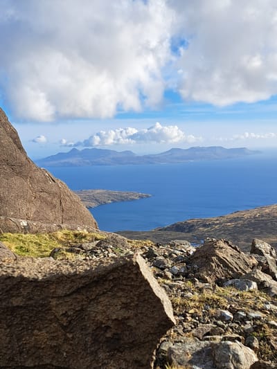 This outdoor shot, taken at Isle of Skye in the United Kingdom, captures a dramatic landscape featuring a craggy terrain. The foreground is dominated by large, textured rock formations with patches of green moss and grass clinging to the surface. The image is oriented vertically, placing the sky and sea along the horizontal axis. The sky is a blend of blue and white, with puffy clouds scattered across the horizon. Below the sky, a body of water stretches into the distance, framed by mountainous landmasses on either side. A slender, pointed rock formation rises from the water, adding to the scene's rugged beauty.