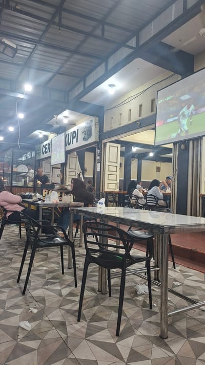 The image depicts a busy outdoor seating area of a restaurant or cafe, potentially located in the city of Keuniree, Indonesia. The scene is filled with people seated at tables, engaged in conversations and enjoying their meals. Several tables are occupied by individuals or groups, with food and drinks visible. The environment appears to be somewhat cluttered, with napkins and wrappers scattered on the floor, suggesting a recent influx of customers. In the background, there is a screen displaying a sports event, likely a soccer game, indicating a communal viewing experience. The architecture suggests a casual dining establishment with an open-air setup.