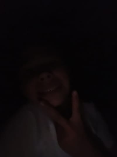 The image depicts a low-light, close-up selfie of an individual making a peace sign with their right hand. The person's face is partially visible, with their mouth open, suggesting they might be speaking or laughing. The background is dark and indistinct, offering no clues about the location. There is no information within the image that can place it in Opoczno, Poland, or any other specific city.