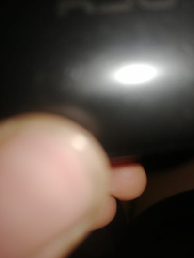 The image depicts a close-up, out-of-focus view of a human finger in the foreground, appearing to be touching or pointing towards a dark surface illuminated by a bright, diffused light source. The background is dominated by this dark surface, likely a device or object, with the light creating a halo effect. No specific location in Pervomaisk, Ukraine, is identifiable. The focus is extremely shallow, rendering most details of the environment indistinct.