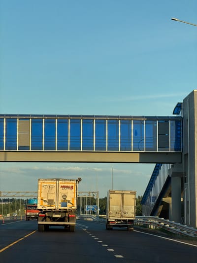 The image shows a highway overpass with a pedestrian walkway, seen from the perspective of a vehicle traveling behind two trucks. The walkway, with its blue glass panels and metal framing, spans across the highway. The sky is clear and blue, with some faint clouds visible in the distance. The highway is paved and has clear lane markings. The foreground shows the rear of two trucks, one white and one light-colored. The white truck has markings that read "MEGA 3 LIGHT" and a license plate that appears to be Russian. The environment is likely a semi-rural or suburban area, given the presence of trees lining the highway in the background. The overpass and highway suggest a well-developed transportation infrastructure. There are no people visible in the image. The scene appears to be captured during daylight, possibly in the late afternoon, due to the lighting and shadows.