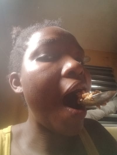 A close-up image depicts an individual with dark skin and dark hair, situated in the city of Iyiowa Odekpa, Nigeria. The subject is viewed from a low angle, with their right eye partially visible and their mouth opened wide. A metallic spoon containing cooked rice and other small food components is positioned at the entrance of the open mouth. The individual is wearing a yellow ribbed garment. The background features a light-colored ceiling, a darker-toned wall segment, and horizontal window blinds or louvers.