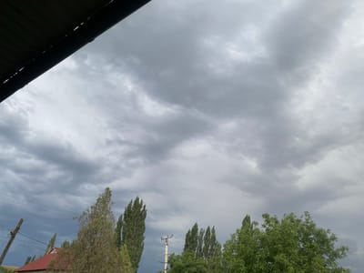 The image depicts an overcast sky filled with dramatic, dark grey clouds, suggesting impending rain or a storm. In the foreground, a dark, corrugated roof juts in from the top left corner, casting a shadow over part of the scene. Below the sky, a row of tall, slender trees with lush green foliage dominates the mid-ground and extends into the right side of the frame. To the left, a traditional house with a reddish-brown roof is partially visible, nestled amongst the trees. A wooden utility pole with wires crisscrossing is also present, hinting at a rural or semi-urban setting. The overall atmosphere is somber and slightly ominous, amplified by the heavy cloud cover and the muted natural light. The location cues, such as the architecture and the type of trees, are consistent with a rural setting in Kyrgyzstan. The time of day is difficult to ascertain precisely due to the cloud cover, but the lighting suggests it could be late afternoon or early evening. There is no visible text in the image.