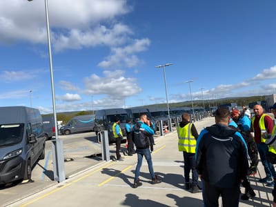 The image depicts an outdoor scene at what appears to be an Amazon logistics facility in Friedrichsdorf, Germany, under a bright, clear blue sky with scattered white clouds. The main subjects are numerous dark grey Amazon Prime delivery vans and a group of approximately ten to fifteen men, presumably Amazon delivery drivers or staff.
The vans are neatly parked in rows within a large, paved area, marked with yellow and blue lines. Each van prominently features the "prime" text and the Amazon smile logo on its side. Several vans are positioned near what look like charging stations or securing bollards.
The men are gathered in small groups across the parking lot, casting distinct shadows due to the bright daylight. Many are dressed in Amazon-branded attire, including blue and black jackets with the Amazon smile logo on the back, or high-visibility yellow safety vests. Their activity suggests they are either preparing for a shift, receiving instructions, or awaiting deployment.
In the foreground, a man with his back to the camera wears a black and blue Amazon jacket. Another man in a similar jacket stands in the mid-ground. Tall, slender lampposts with modern light fixtures are visible throughout the area. In the background, rolling hills covered with trees provide a natural backdrop to the industrial setting. A license plate on one of the distant vans appears to begin with "M-AZ".