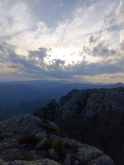A high-angle shot captures a mountainous landscape under a cloudy sky. In the foreground, rocky formations with moss and sparse vegetation dominate the lower portion of the frame. Behind the rocks, a series of mountain ranges fade into the distance, their peaks and valleys softened by atmospheric haze. The sky is filled with a mix of gray and white clouds, with the sun partially obscured, casting a diffused light across the scene. The overall composition suggests a remote, elevated location. It is identified as Ichera, Bulgaria.