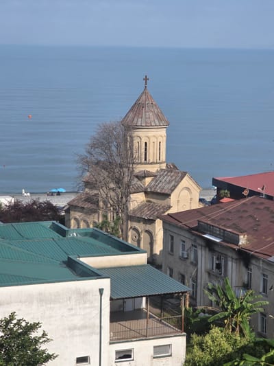 This is a high-angle, eye-level, full shot photograph of a coastal city scene in Sarpi, Georgia. In the foreground, the tops of several buildings are visible, featuring green and brown tiled roofs and white walls. One building has a balcony with a railing. In the mid-ground, a large, tan-colored church with a domed roof and a cross on top stands prominently. The church has arched windows and decorative details. Next to the church are more buildings with brown tiled roofs, some appearing older and in a state of disrepair. A bare-branched tree is situated in front of the church. Beyond the buildings and the church, the sea stretches to the horizon, a calm expanse of blue water. A small orange buoy is visible in the water to the left. The sky is clear and blue, with a slight haze near the horizon. A beach area with two blue umbrellas and a few figures can be seen along the coastline. The overall scene depicts a coastal urban environment with a notable religious structure overlooking the sea.
