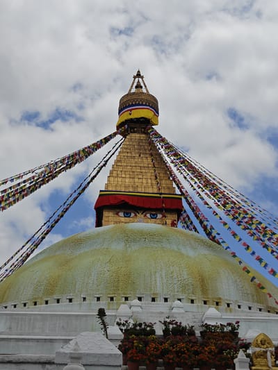This image shows the Boudhanath Stupa in Gokarneshwar, Nepal, a UNESCO World Heritage site. The stupa is a large dome with a golden spire topped with a canopy. Prayer flags in various colors are strung from the spire, fluttering in the sky. The eyes of Buddha are painted on the sides of the spire. The base of the stupa is a white circular structure with multiple levels. In the foreground, potted plants with colorful flowers are arranged. The sky above is cloudy with patches of blue.
