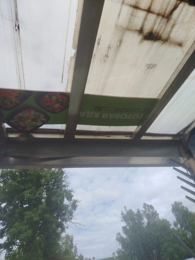 This media file shows an outdoor scene, looking upwards from underneath a covered structure. The structure appears to be made of translucent corrugated plastic panels supported by metal beams. The plastic panels are dirty and stained, with some damage visible.
Attached to the underside of the structure is a green banner displaying images of food in square containers. Text on the banner reads "ГОТОВАЯ ЕДА С СОБОЙ" which translates from Russian to "READY MEAL TO GO".
Below the covered structure, a view of trees and a cloudy sky is visible. The trees are lush and green, suggesting it is summertime. The sky is overcast, indicating a possibly humid or rainy day.
The scene appears to be in an outdoor public area, possibly a market, food stall, or an entrance to a building with an awning. There are no people visible in the image. The overall impression is one of a slightly weathered but functional outdoor setup.