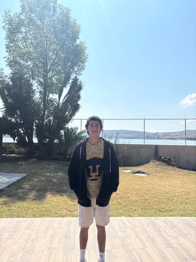 The photo shows a young man standing outdoors. He is wearing a black jacket over a patterned shirt, white shorts, and white socks with a black Nike logo. He also has a hat or cap on his head.
The background consists of a landscape featuring a body of water visible through a fence. There is green grass to one side and tiled flooring to the other. A tree is visible on the left.
The image was most likely taken in El Oasis Valsequillo, Mexico.