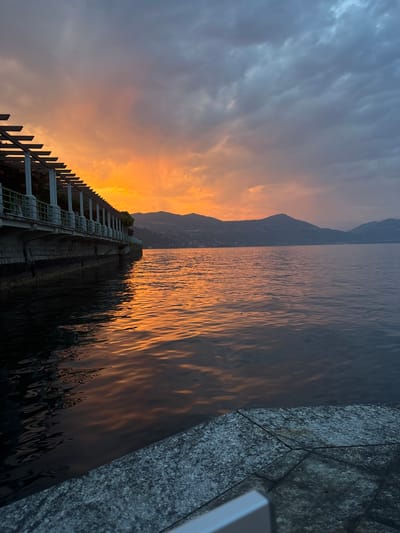 This is a vertical, medium shot of a lakefront promenade at sunset in Arona, Italy. The sky is filled with dramatic clouds, transitioning from a fiery orange and yellow near the horizon to a deep grey and blue overhead. The setting sun casts a warm, golden reflection on the surface of the lake, which stretches out towards a distant, silhouetted mountain range.

In the foreground, a stone-tiled walkway forms a diagonal line from the lower right corner. To the left, a stone retaining wall supports a promenade lined with a white pergola structure, supported by stone pillars. The pergola's beams cast shadows on the walkway beneath. The lake laps gently against the retaining wall. The overall atmosphere is serene and picturesque, typical of a lakeside sunset view. There are no people visible in the image.